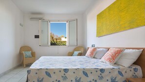 Een slaapkamer met een bed, twee stoelen, een raam en een gele afbeelding aan de muur.