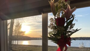 Ett bord med en vas med blommor, en presentförpackning och utspridda rosenblad, placerat vid ett fönster med utsikt över en sjö i solnedgången.