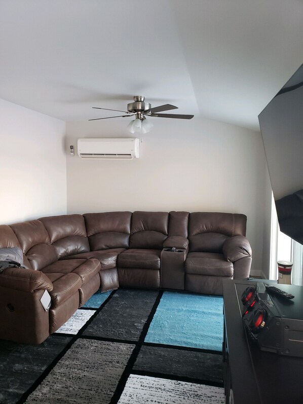 A living room with a brown leather sectional sofa, a ceiling fan, an air conditioning unit, a blue and black rug, and a television on a stand. - Tranquility on Rogers Pond (Stanwood)