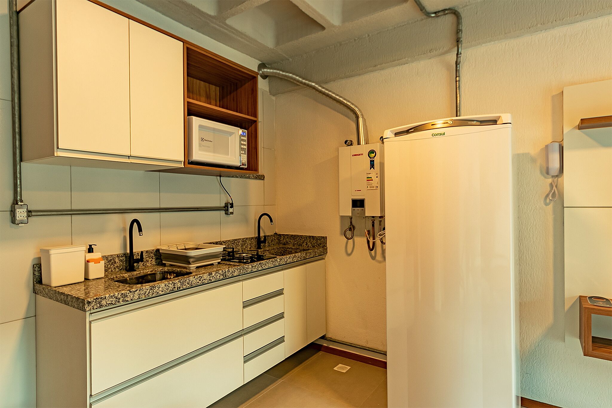 Cozinha com armários brancos, bancada de granito, micro-ondas embutido, aquecedor de água e geladeira.
