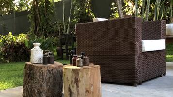 Um espaço de jardim com um sofá de vime e dois banquinhos de madeira, cercado por vegetação e árvores.