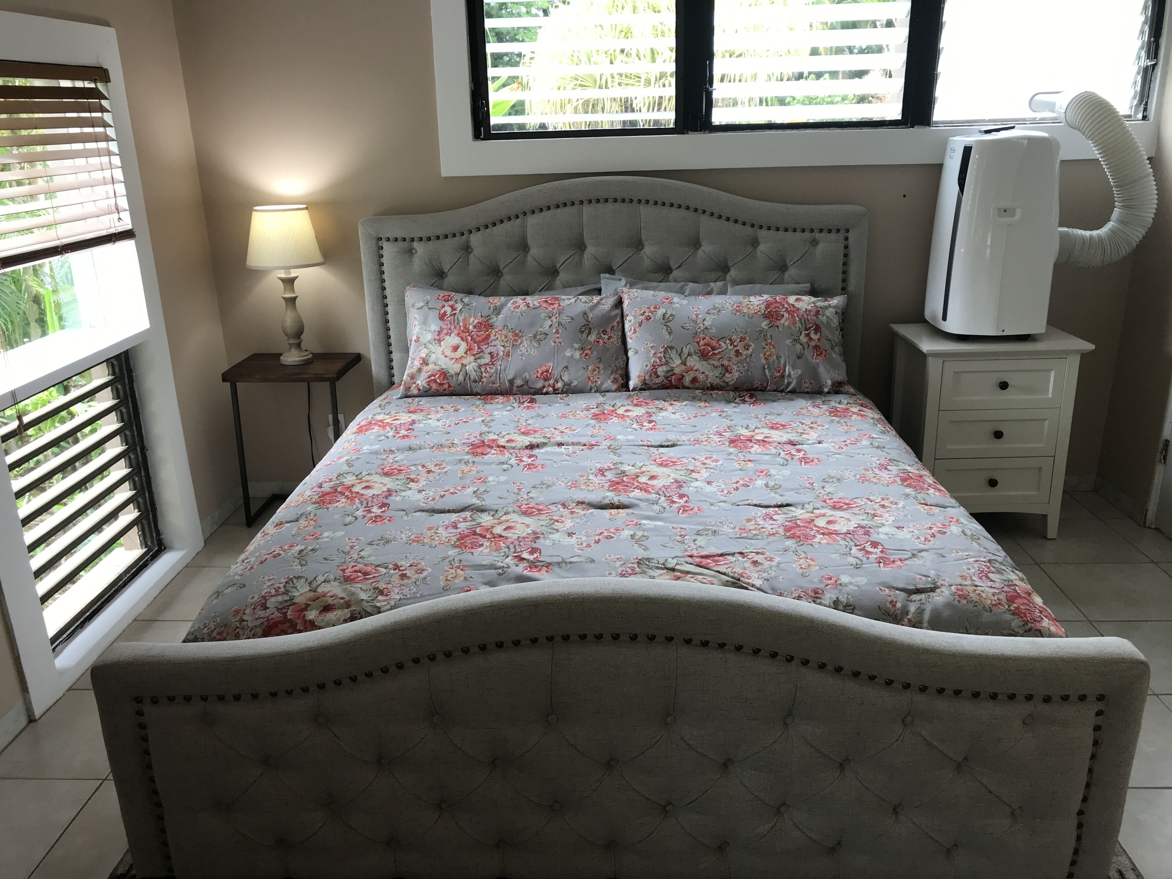 Een slaapkamer met een bed, een nachtkastje, een lamp, een raam met zonwering en een airconditioning.