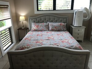 Ein Schlafzimmer mit Bett, Nachttisch, Lampe, einem Fenster mit Jalousien und einer Klimaanlage.