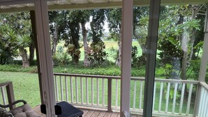 A covered porch with a wicker chair and a small table, overlooking a grassy yard with trees and a fence.