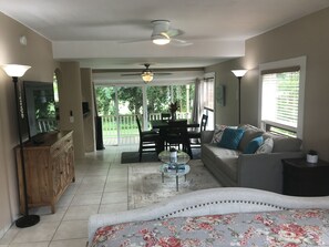 Ein geräumiges Wohnzimmer mit einer Couch, einem Couchtisch und einem Fernseher auf einer Holzkommode. Ein Balkon mit Esstisch und Stühlen sowie ein Deckenventilator sind ebenfalls vorhanden.