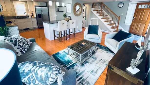 A spacious living room with a grey sofa, a coffee table, and a TV stand. A kitchen with white cabinets and a dining area with a white table and chairs are visible in the background. - Coastal Cottage - Porch, Grill, Walk to Portsmouth (Kittery)
