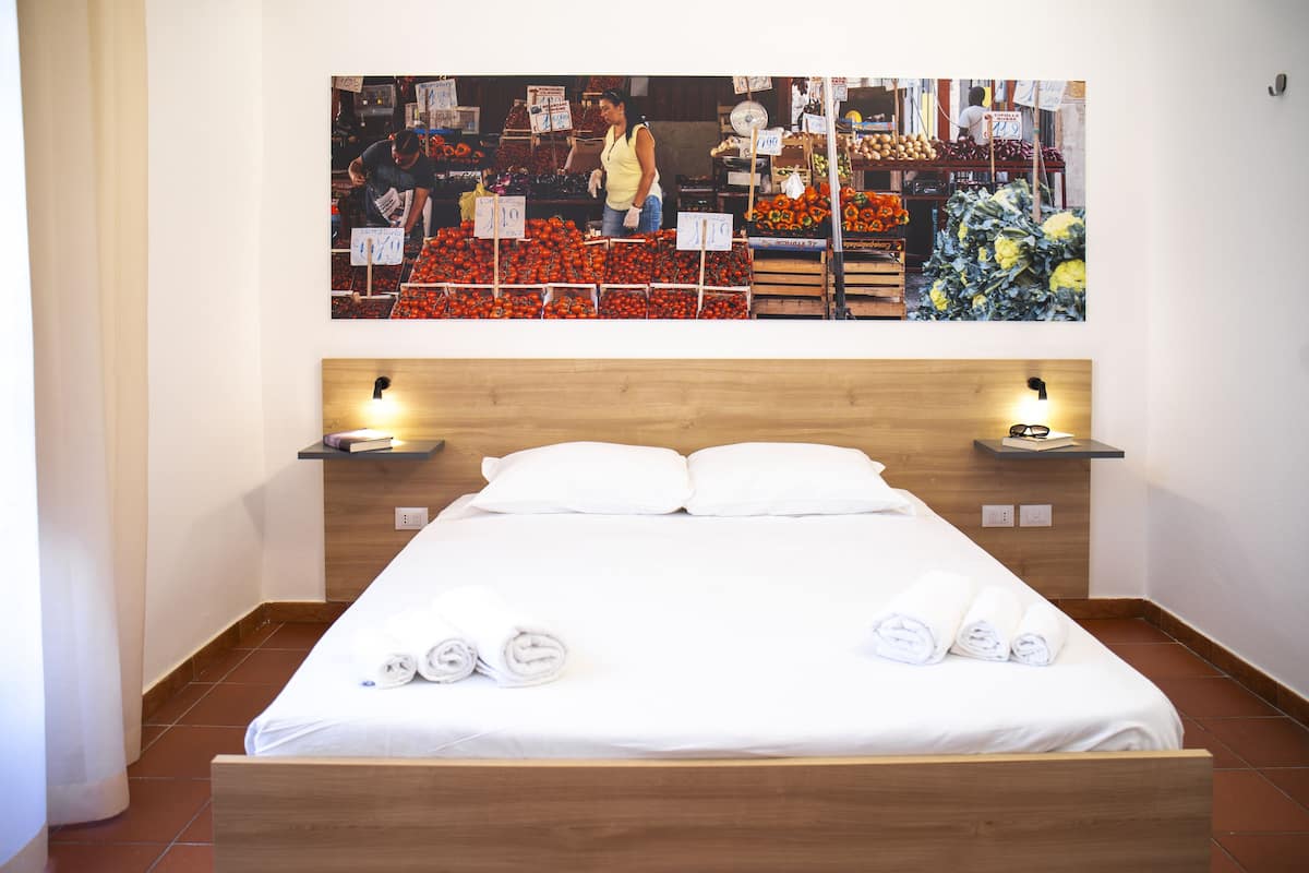 A hotel room with a large bed, two bedside tables, and a wall-mounted artwork depicting a market scene.