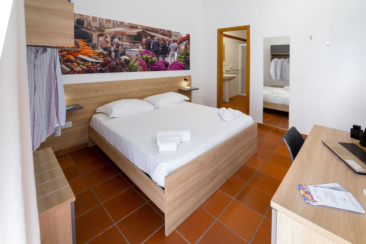 A hotel room with a wooden bed, a desk with a laptop, a bathroom with a sink and mirror, and a large painting on the wall.