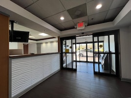 Un área de recepción con un mostrador de madera, una pared blanca con textura y una puerta de cristal que da al exterior.