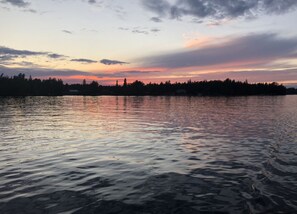 Un lac paisible au coucher du soleil, avec la silhouette d’arbres et de bâtiments sur la rive opposée.