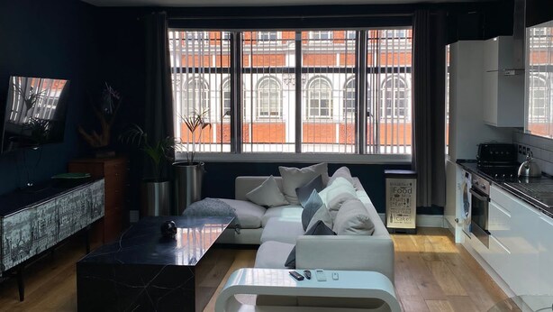 A modern living room with a sofa, a coffee table, and a TV. There is a skylight above the sofa and a kitchen area with a stove and sink.