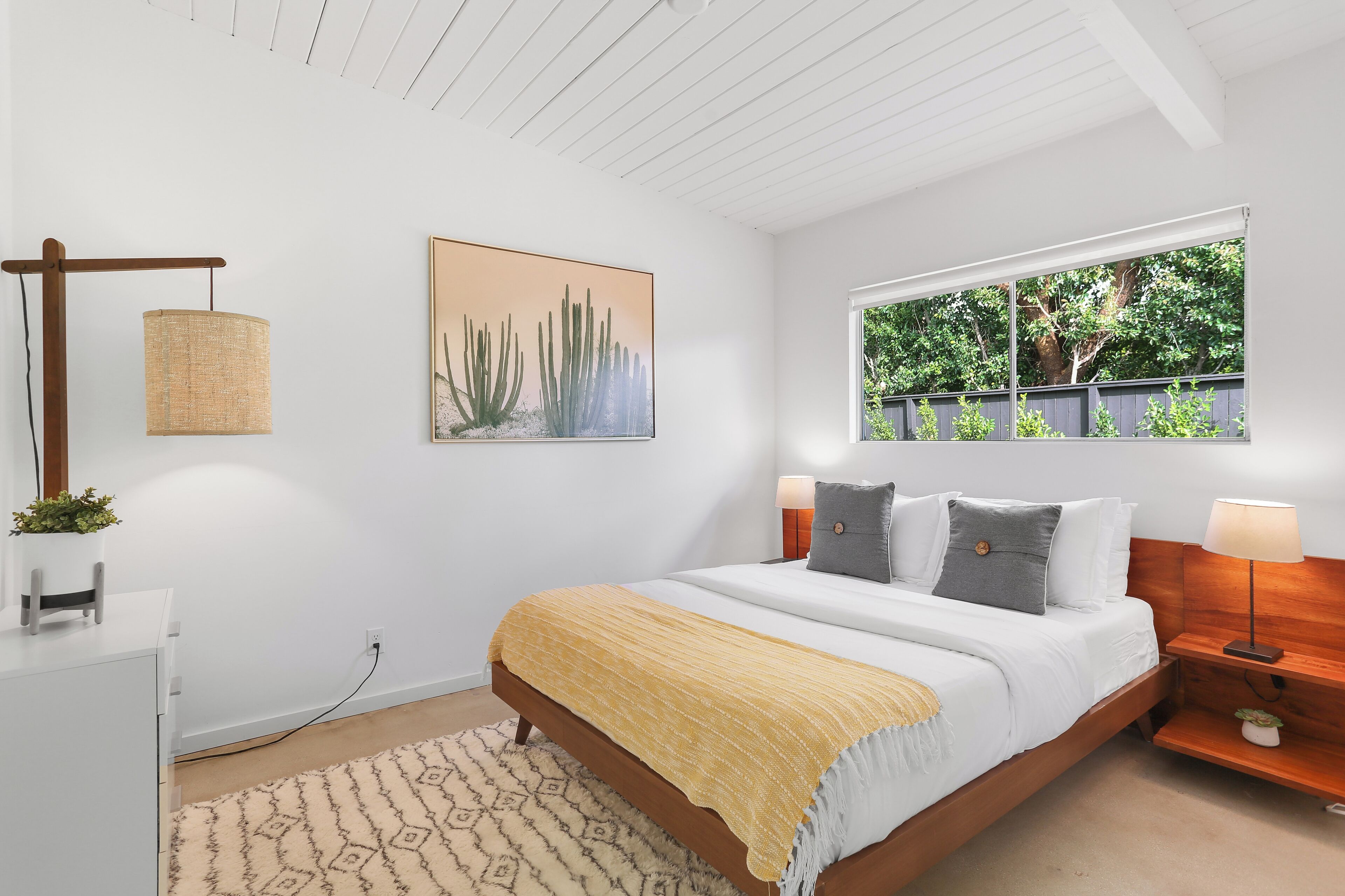 A modern bedroom with a wooden bed, a bedside table with a lamp, a wall-mounted lamp, a small plant, and a window with a view of greenery.