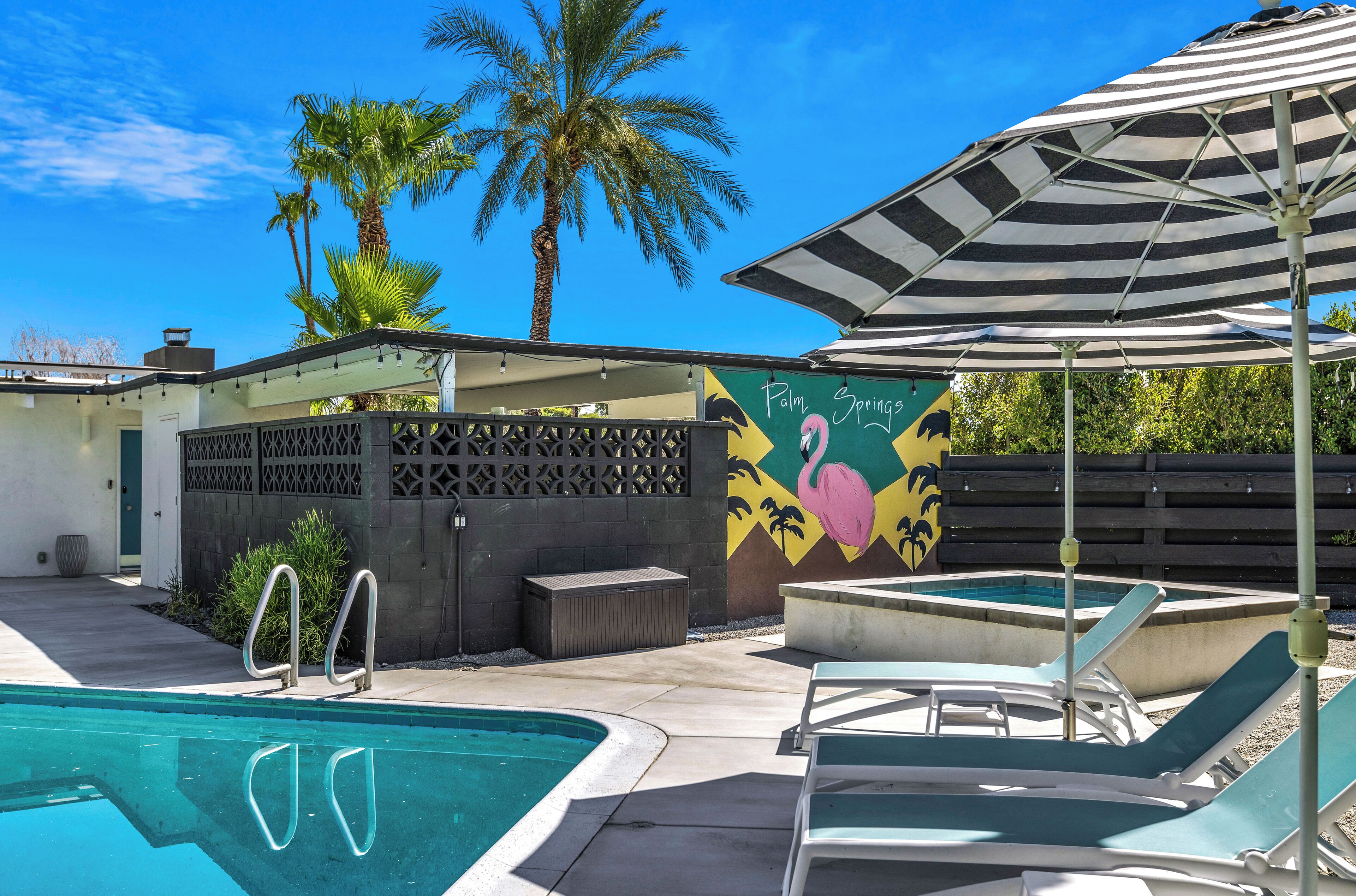 A swimming pool with lounge chairs and a striped umbrella, surrounded by palm trees and a mural.