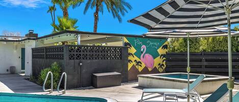 A swimming pool with lounge chairs and a striped umbrella, surrounded by palm trees and a mural.