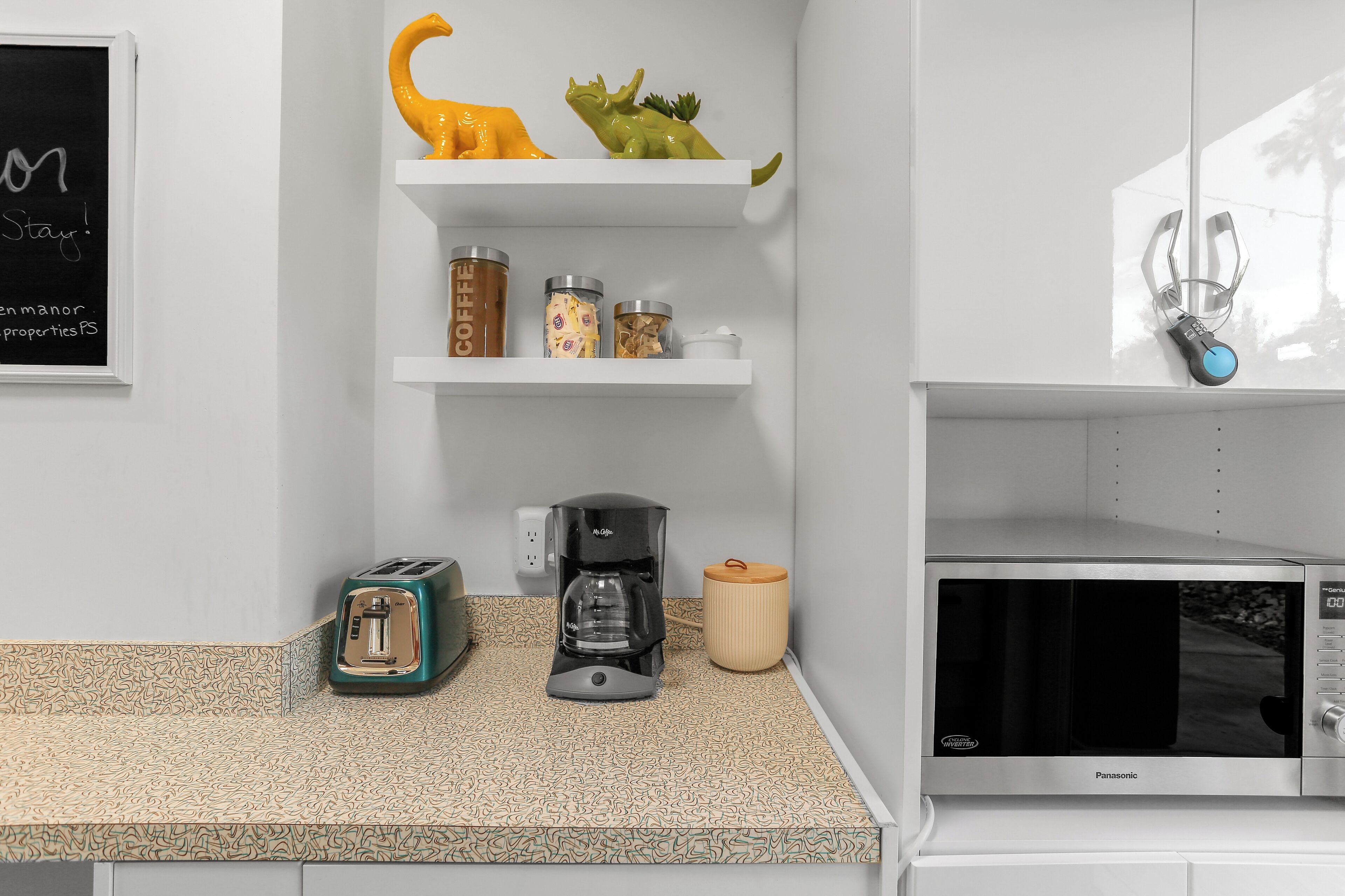 Cozinha com torradeira, máquina de café e micro-ondas. Há prateleiras com objetos decorativos e recipientes.