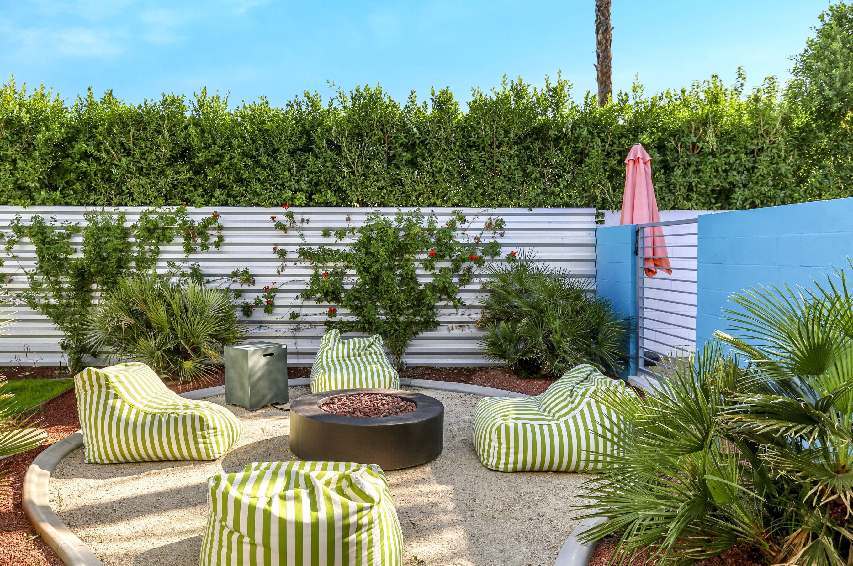 A patio area with green-striped bean bags, a circular seating area with a fire pit, and a blue wall with climbing plants.