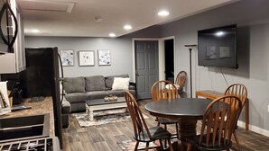 A modern living room with a gray sofa, a dining table with wooden chairs, a flat-screen TV mounted on the wall, and a kitchen area with a refrigerator and cabinets. - Country, close to town, sled trails, skiing, large snowmobile boat trailer space (Sault Ste. Marie)