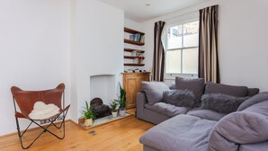 A living room with a grey sofa, a brown leather chair, a bookshelf, and a window with curtains. - Charming & Peaceful 1BD Home in Camden Town (London)