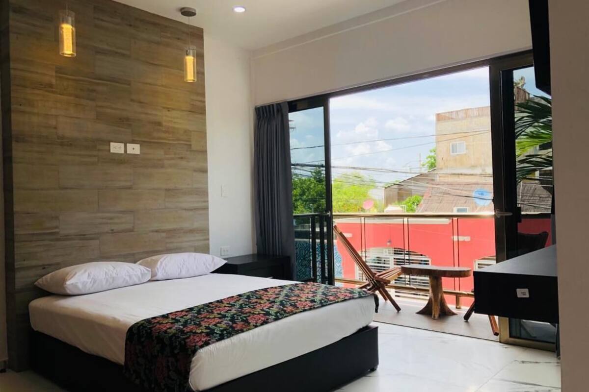 A bedroom with a large bed, a wooden accent wall, a balcony with a table and chair, and a view of the outside.