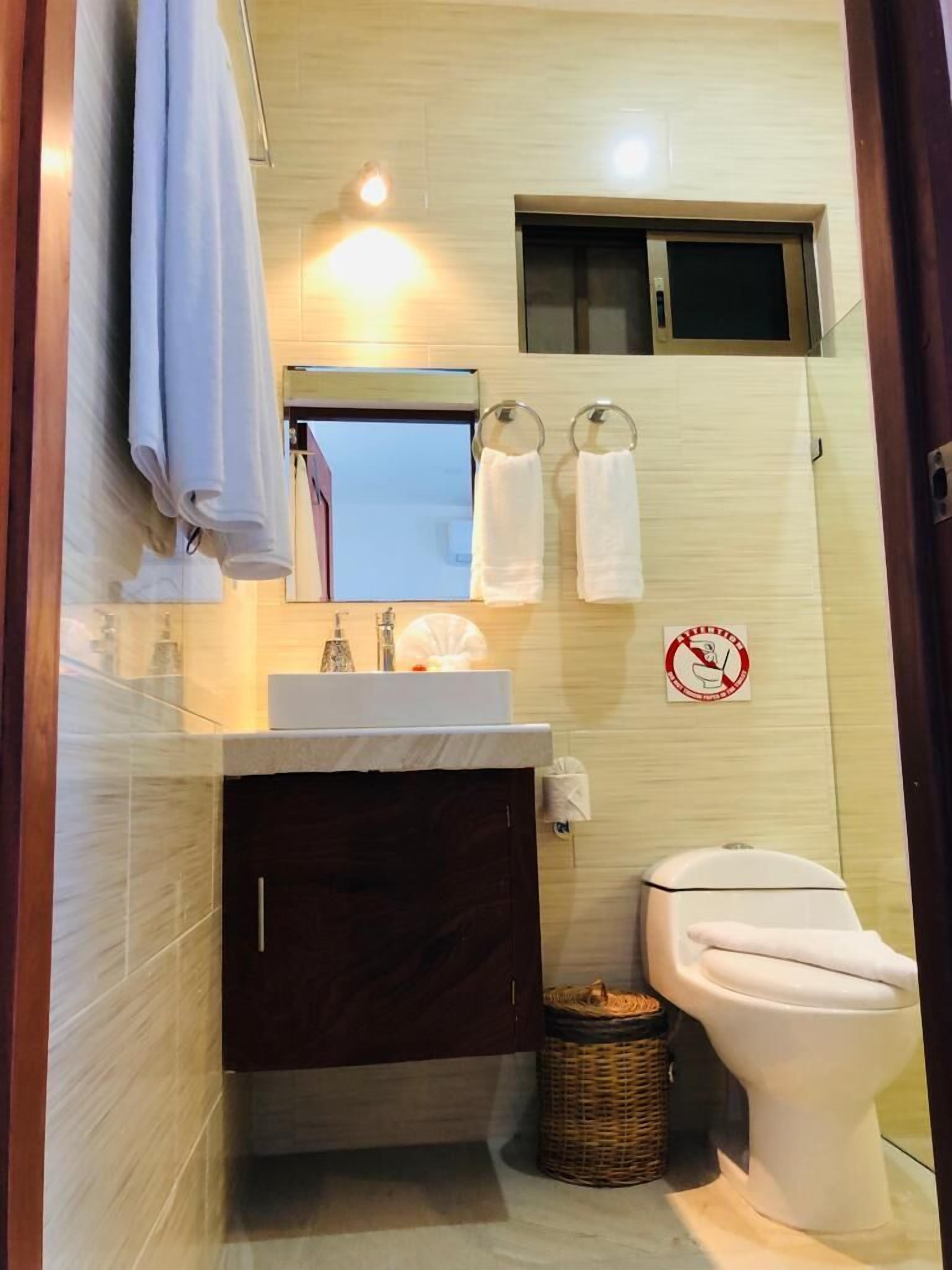 A compact bathroom with a sink, mirror, and toilet. There are two towels hanging on hooks, a trash bin, and a no-smoking sign on the wall.