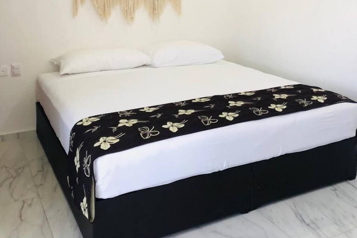 A bedroom with a bed, white walls, a macrame wall hanging, and a ceiling-mounted air conditioner.