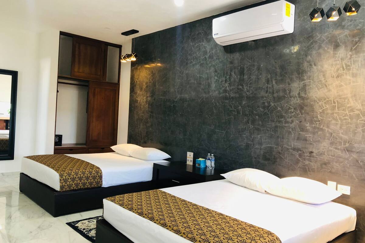 A modern hotel room with two beds, a dark textured wall, an air conditioning unit, and a small table with water bottles.
