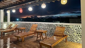 Un área de estar al aire libre con tumbonas de madera y una pared estampada, con vistas a un paisaje urbano al atardecer.
