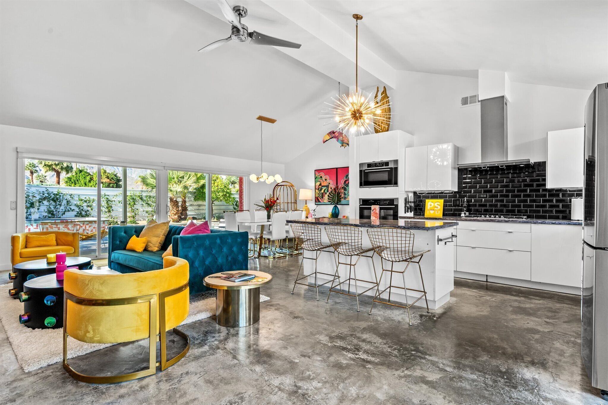 A modern kitchen with white cabinets, a black backsplash, and stainless steel appliances. There is a bar area with bar stools and a dining table with chairs. The living area has a blue sofa, yellow armchairs, and a coffee table.