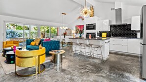 A modern kitchen with white cabinets, a black backsplash, and stainless steel appliances. There is a bar area with bar stools and a dining table with chairs. The living area has a blue sofa, yellow armchairs, and a coffee table. - Birdcage Palm Springs - A Ryson Property (Palm Springs)