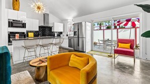 A modern kitchen with white cabinets, a stainless steel refrigerator, and a dining area with a yellow sofa and metal chairs. - Birdcage Palm Springs - A Ryson Property (Palm Springs)