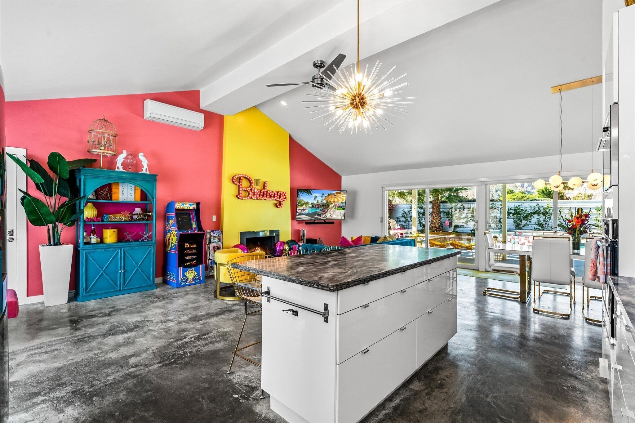 A modern kitchen with a central island, a colorful wall with a neon sign, and a large window offering a view of the outdoors.