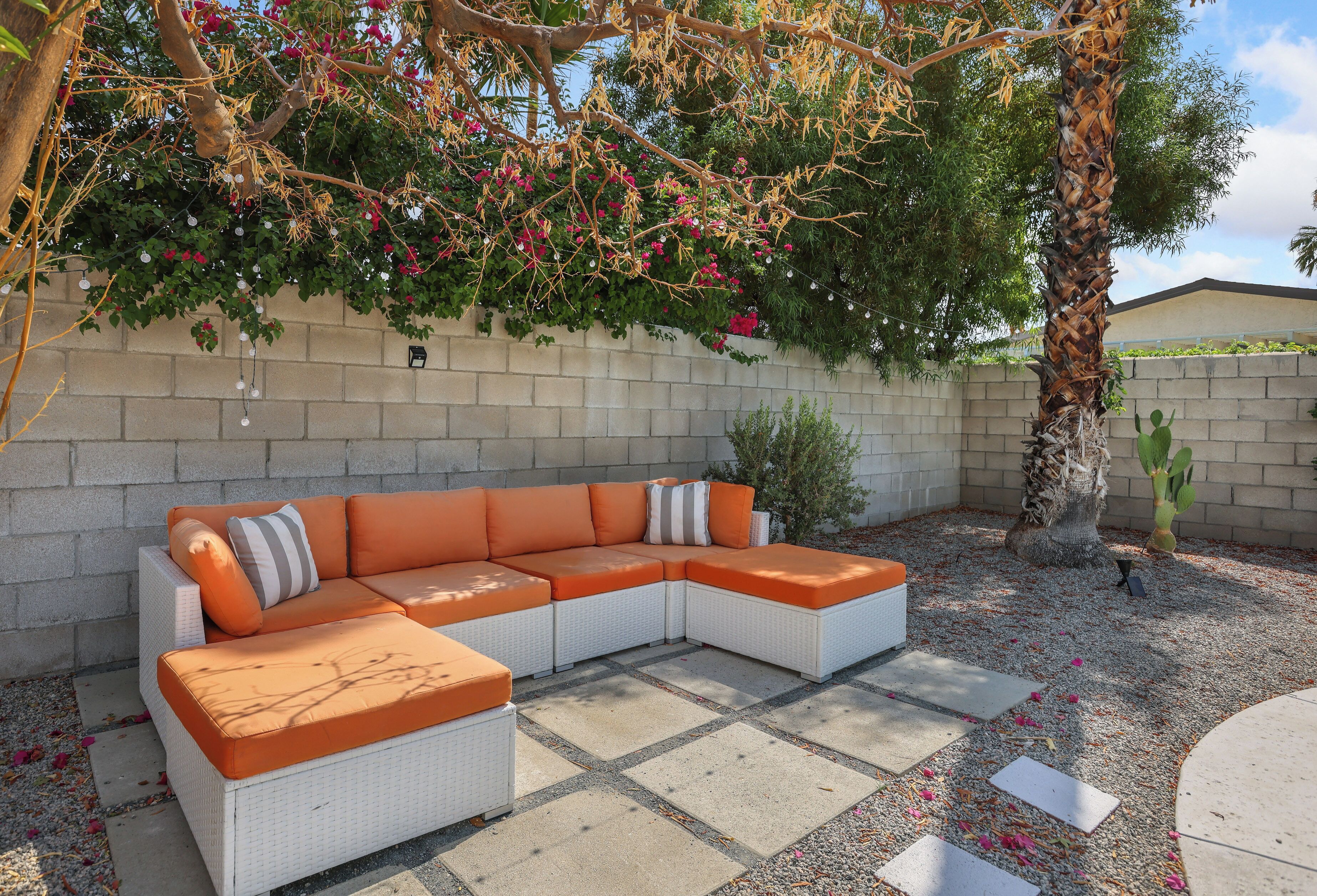 Ein modulares Sofa mit orangefarbenen Kissen vor einer beigen Wand, umgeben von einer Schotterterrasse und einem Baum mit rosa Blüten.