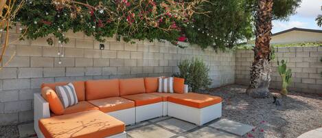 Ein modulares Sofa mit orangefarbenen Kissen vor einer beigen Wand, umgeben von einer Schotterterrasse und einem Baum mit rosa Blüten.
