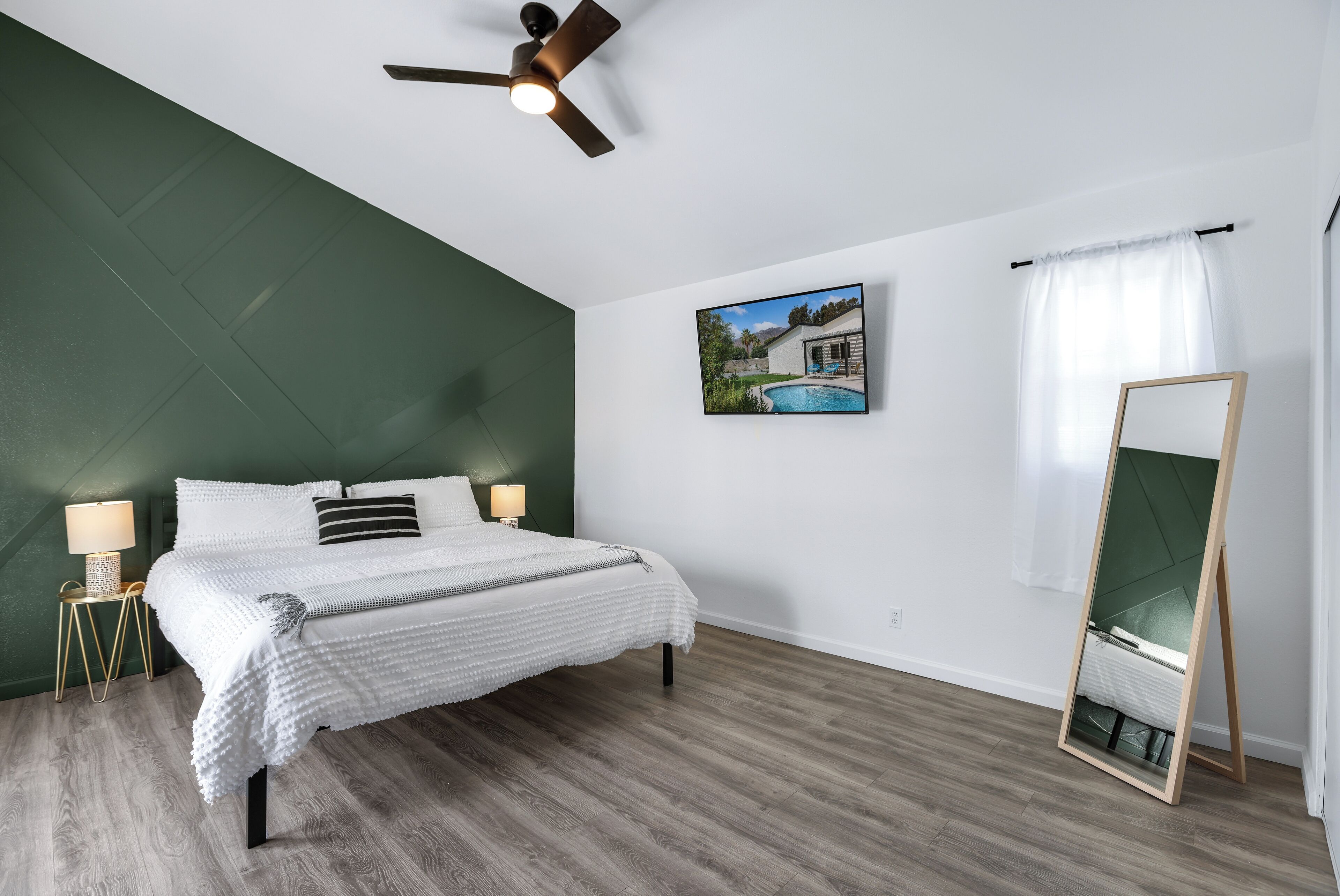 Une chambre à coucher comprenant un lit, un ventilateur de plafond, une table de chevet avec une lampe, un téléviseur fixé au mur et un grand miroir.