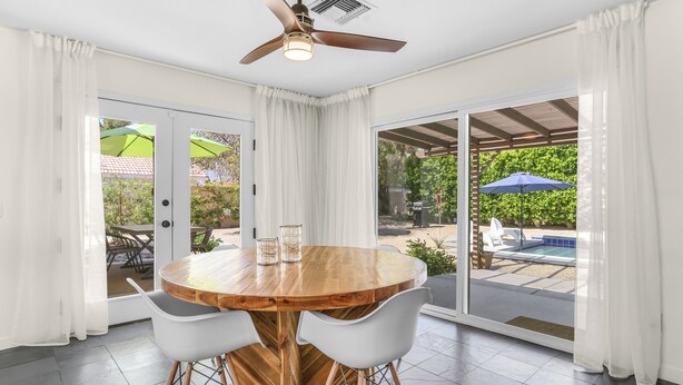 A dining area with a round wooden table and white chairs, large sliding glass doors leading to an outdoor patio with umbrellas. - Tangerine Desert Dreams - A Ryson Property (Palm Springs)