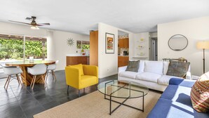 A modern living room with a yellow armchair, a glass coffee table, a white sofa, and a blue sofa. There is a dining area with a wooden table and chairs, and a kitchen in the background. - Tangerine Desert Dreams - A Ryson Property (Palm Springs)