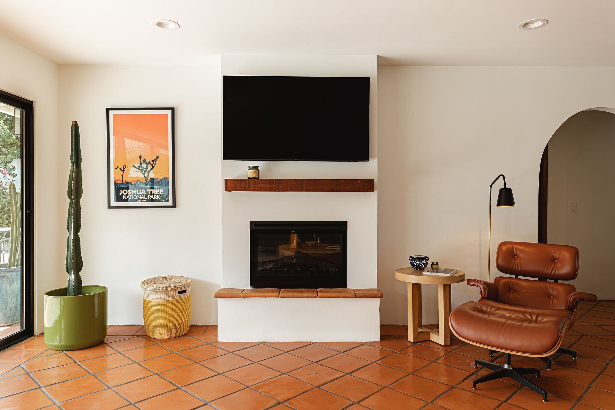 A living room with a flat-screen TV mounted above a fireplace, a cactus in a green pot, a woven basket, and a leather armchair.