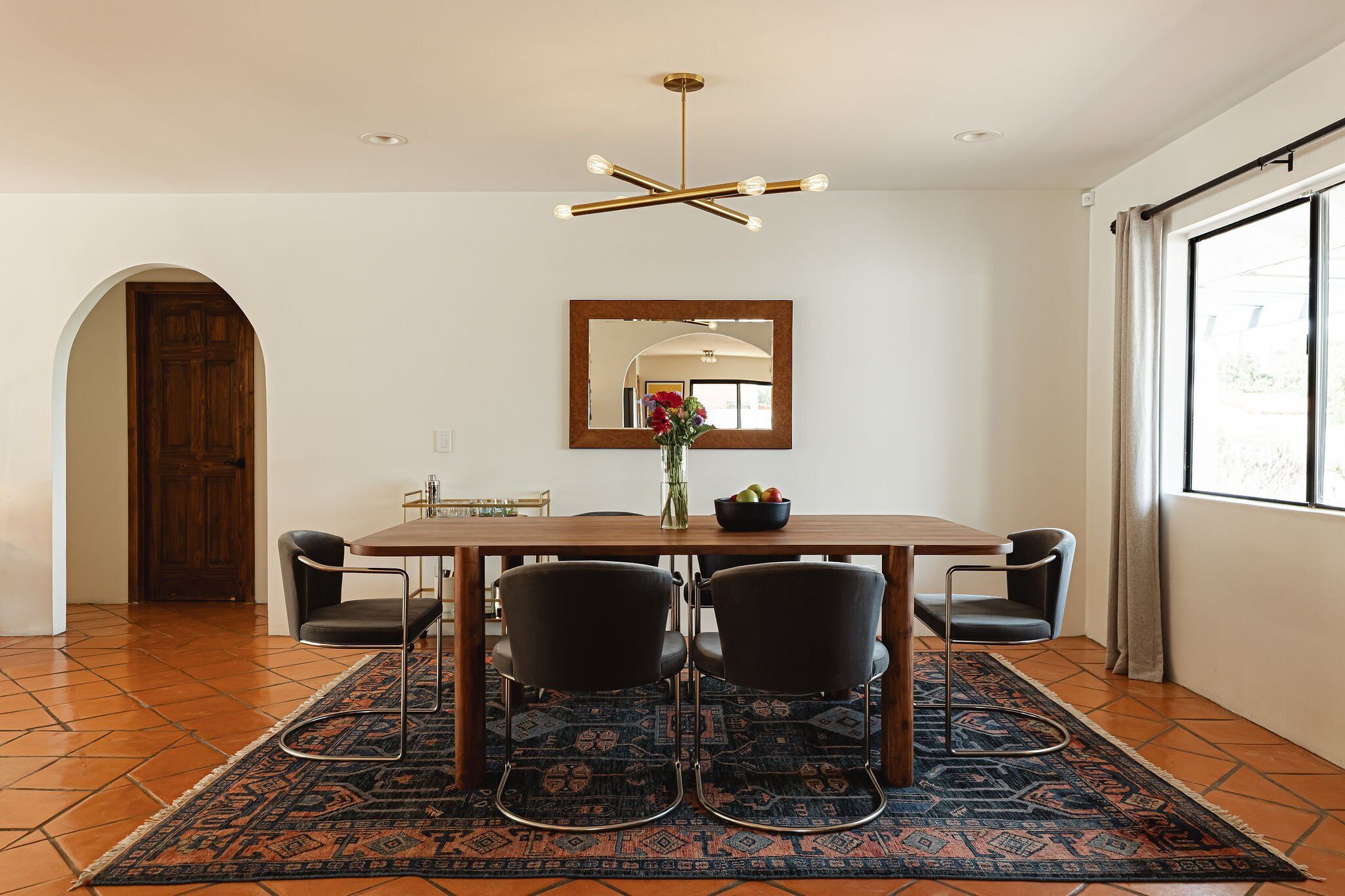 A dining area with a wooden table, four chairs, a vase with flowers, and a large mirror on the wall.