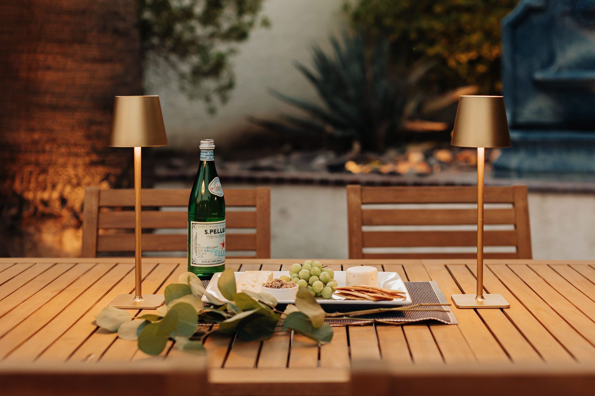 A wooden table with a bottle of S. Pellegrino water, a cheese platter, and a small bowl of nuts. Two lamps with gold shades are on either side of the table.