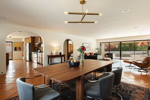 A spacious dining area with a wooden table and chairs, a modern chandelier, and a view of the outdoors through large windows. - Casa Coyote - A Ryson Property (Palm Springs)