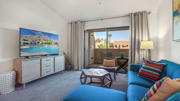 A hotel room with a TV, a sofa, a small table, and a balcony with a view of palm trees and buildings. - Convenient & Cozy Downtown - A Ryson Property (Palm Springs)