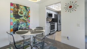 A modern kitchen with stainless steel appliances, a dining area with a glass table and chairs, and a colorful abstract painting on the wall. - Convenient & Cozy Downtown - A Ryson Property (Palm Springs)