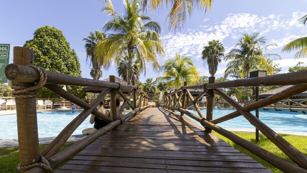 A wooden deck leading to a pool area with palm trees and lounge chairs. - Apart Hotel Le Jardin Caldas Novas (Caldas Novas)