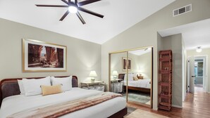 Une chambre d’hôtel avec un grand lit, des tables de chevet avec des lampes, un ventilateur de plafond, un tableau encadré au mur, un miroir et un plancher en bois.