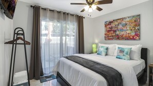 A bedroom with a bed, a ceiling fan, a window with curtains, a wall-mounted TV, and a colorful artwork. - Simple Elegance - A Ryson Property (Indio)
