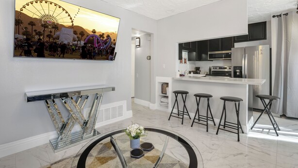A modern kitchen with a central island, bar stools, and a large TV displaying a sunset scene. - Simple Elegance - A Ryson Property (Indio)