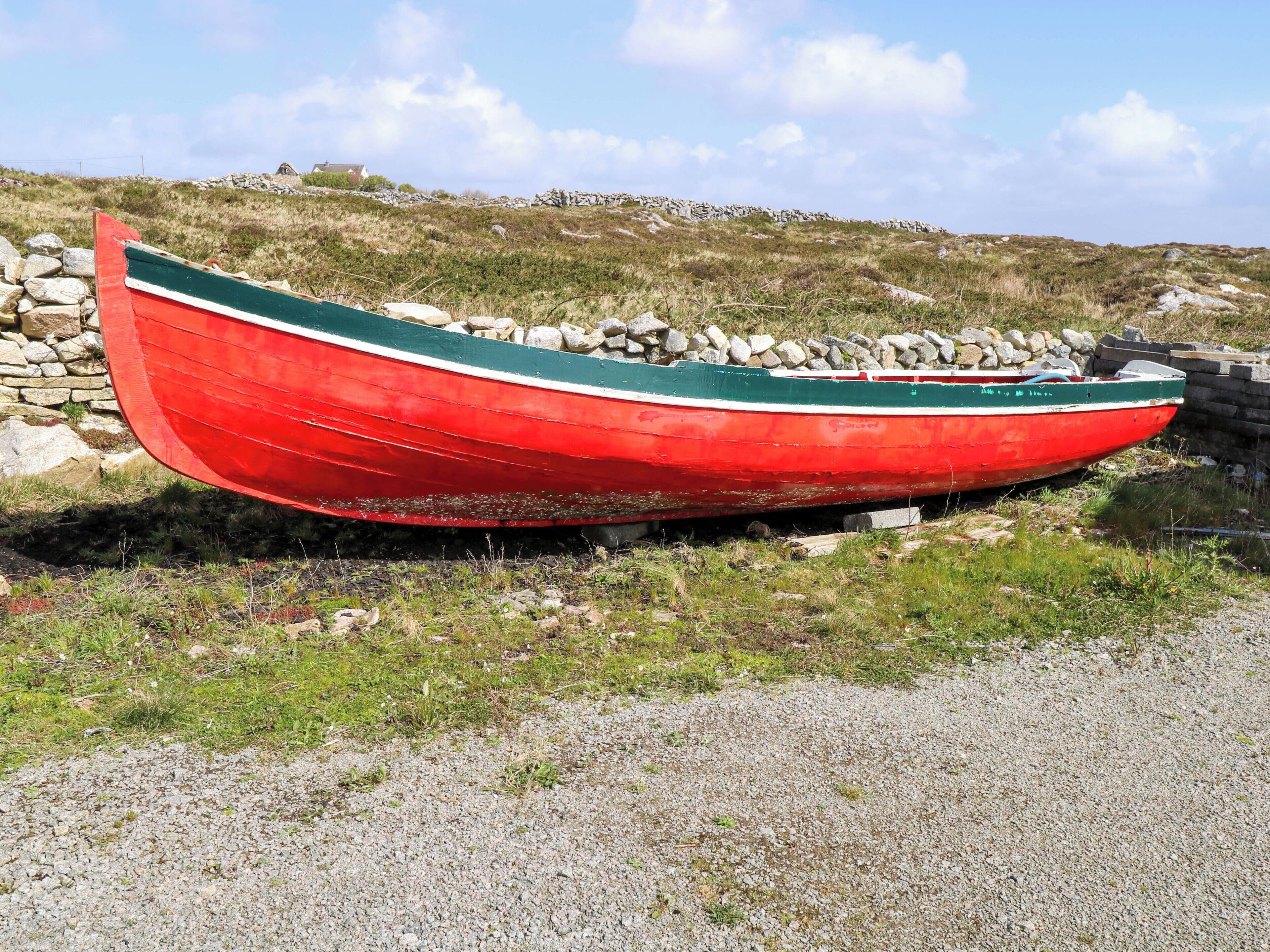 Un barco rojo con una franja verde, reposando sobre un camino de grava con un muro de piedra y una colina cubierta de hierba al fondo.