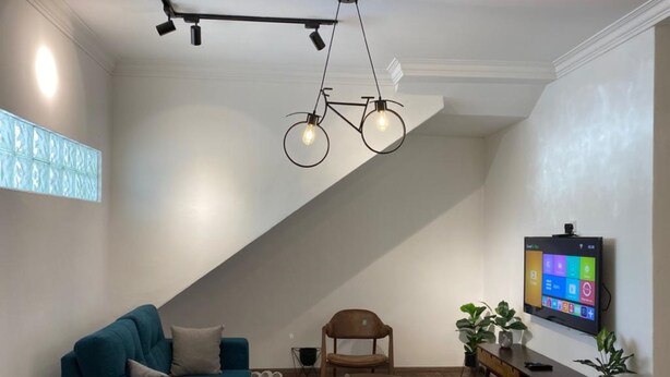 A modern living room with a teal sofa, wooden chairs, a TV on the wall, and a unique bicycle-shaped light fixture.