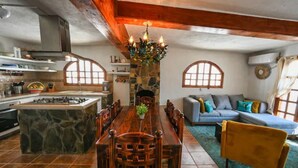 Una cocina amplia con una isla central, mesa de comedor de madera y sillas. Cuenta con una chimenea de piedra, un sofá con cojines y una lámpara de araña.