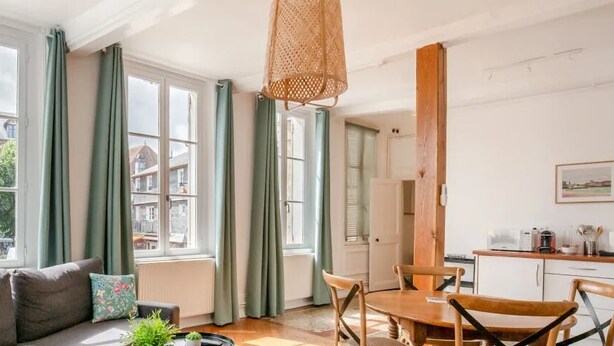 Un salón acogedor con sofá, una mesa de comedor y sillas. Hay ventanas amplias con cortinas, una lámpara colgante y vistas a edificios.
